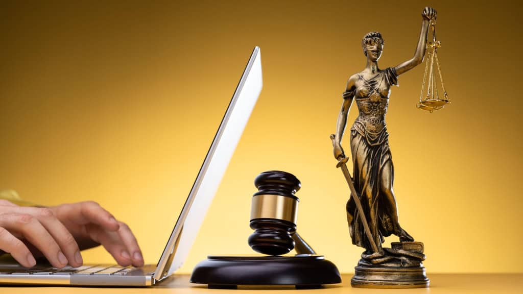 A laptop, a gavel, and the Statue of Justice are displayed on a wooden surface against a golden background, symbolizing legal concepts in the digital age.