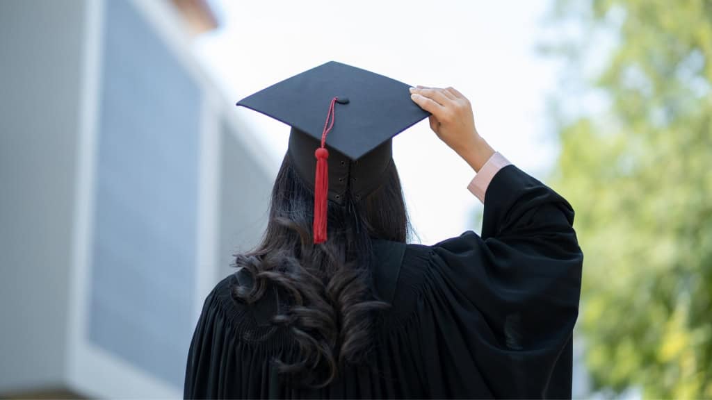 A person wearing a black graduation cap and gown, holding the cap with their hand, with green foliage in the background.