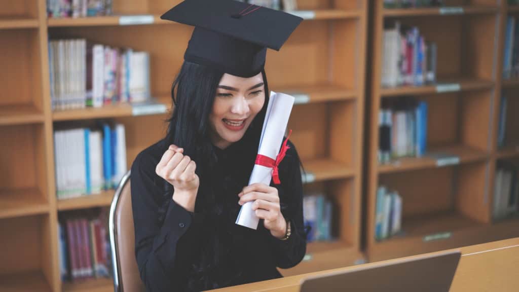 A woman wearing a graduation cap and holding a diploma smiles at a laptop while sitting at a table