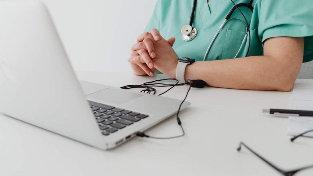A person in scrubs with a stethoscope around their neck sits at a desk with a laptop and glasses.