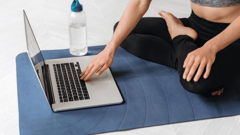 Person sitting on a yoga mat, using a laptop with a water bottle nearby.