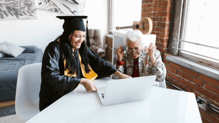 a person in a graduation gown sitting at a table with another person using a laptop