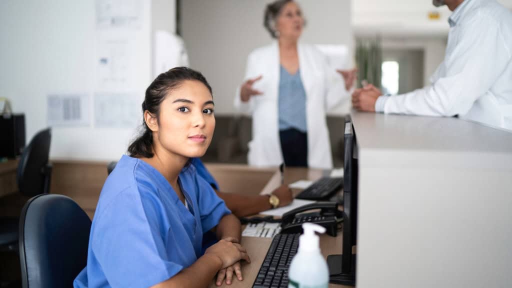 A medical professional in blue scrubs sits at a reception desk with a computer and a bottle of hand sanitizer. In the background, two people in lab coats converse at a counter.