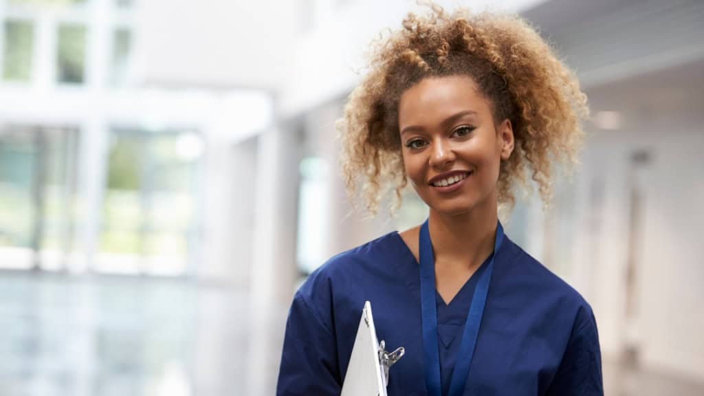 A smiling person in blue scrubs holds a clipboard and wears a lanyard in a hospital hallway.