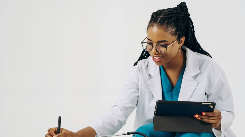 A person wearing a white lab coat and glasses, holding a tablet and writing on a notepad.