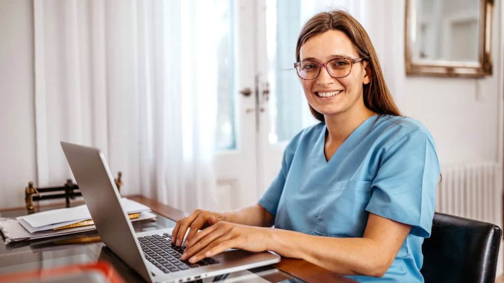 A person wearing blue scrubs and glasses smiles while typing on a laptop at a desk.