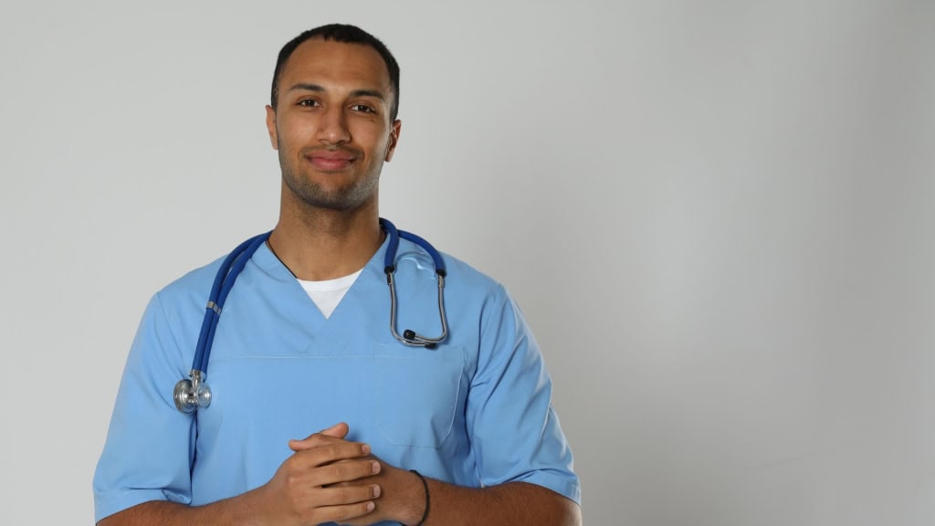 A medical professional wearing light blue scrubs and a stethoscope around their neck, looking towards the camera with clasped hands and a slight smile.
