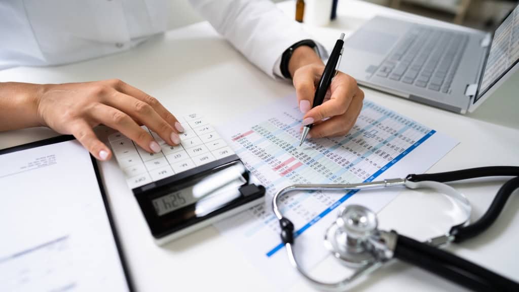 A person in a white coat uses a calculator and pen to review financial documents, with a stethoscope and laptop nearby.