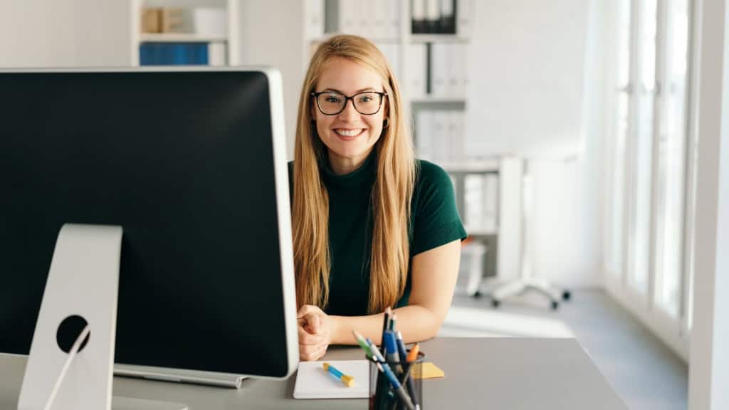Medical Billing and Coding Credentials 2026: CCA, CCS, RHIT & More 1 A smiling person with glasses and long blonde hair wearing a dark green shirt sits at a desk in front of a computer with pens and paper nearby.