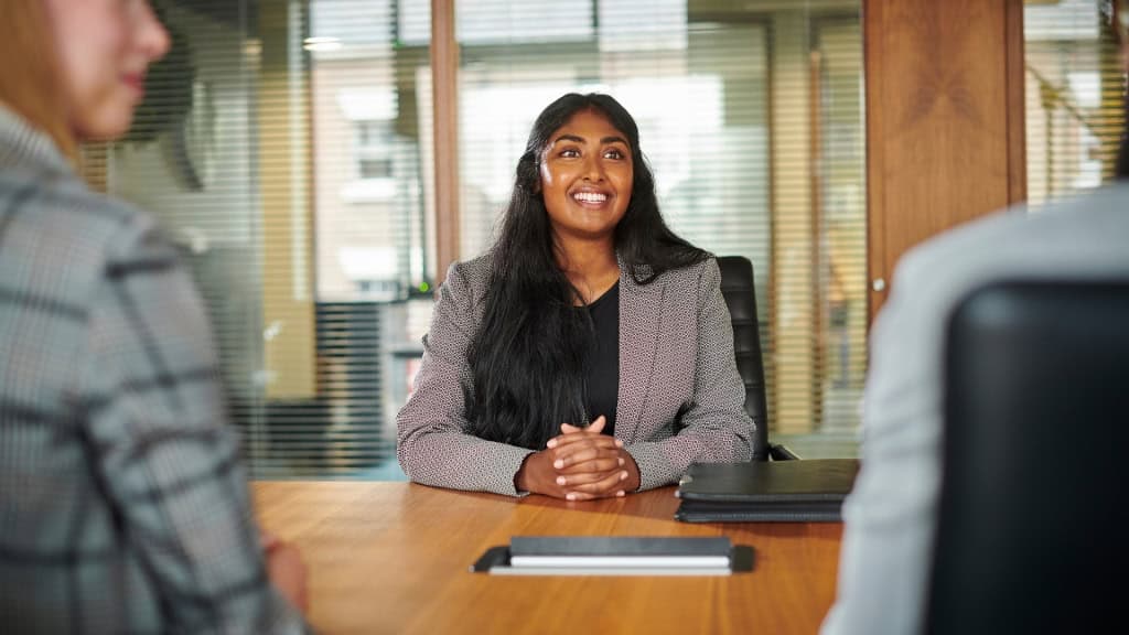 A person in a blazer is sitting at a table with their hands clasped, looking forward and smiling. Two other people are visible in the foreground and background, out of focus.