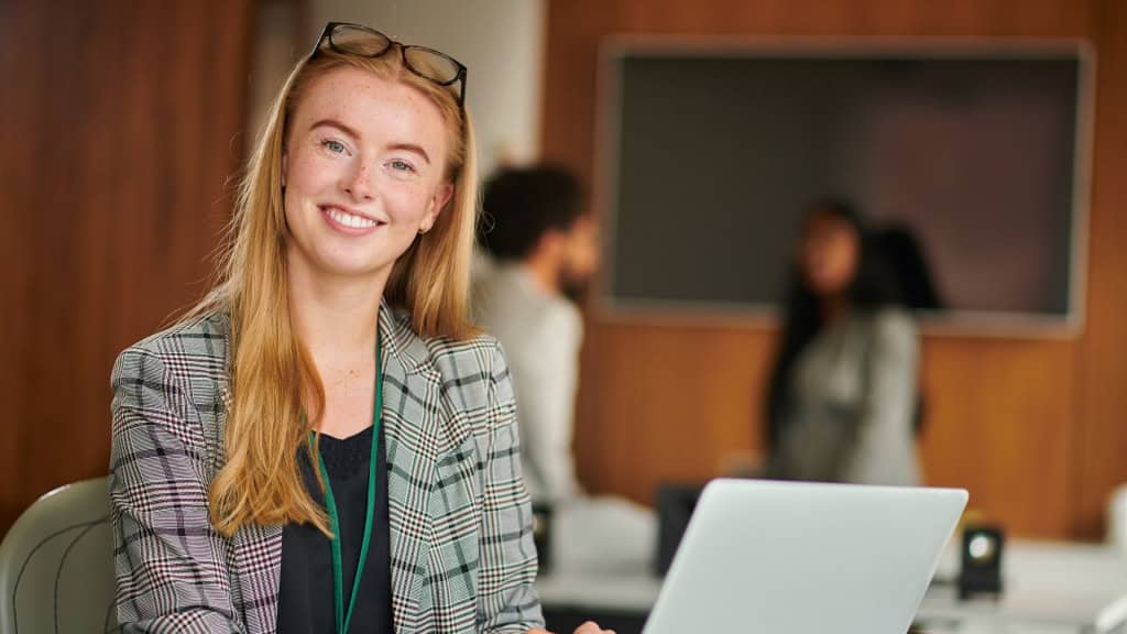 A smiling person with glasses on their head, wearing a plaid blazer and sitting in front of a laptop in an office setting with other people blurred in the background.