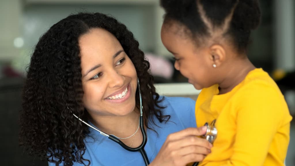 A healthcare professional wearing a blue uniform uses a stethoscope to examine a young child in a yellow shirt.