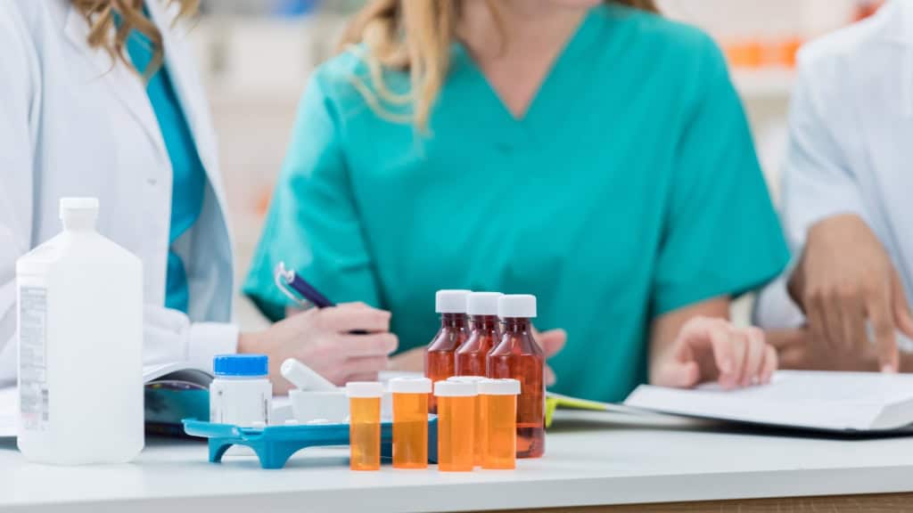 Three individuals in medical attire are gathered around a table with various medication bottles and a large white bottle, one person is writing and another is pointing at a document.