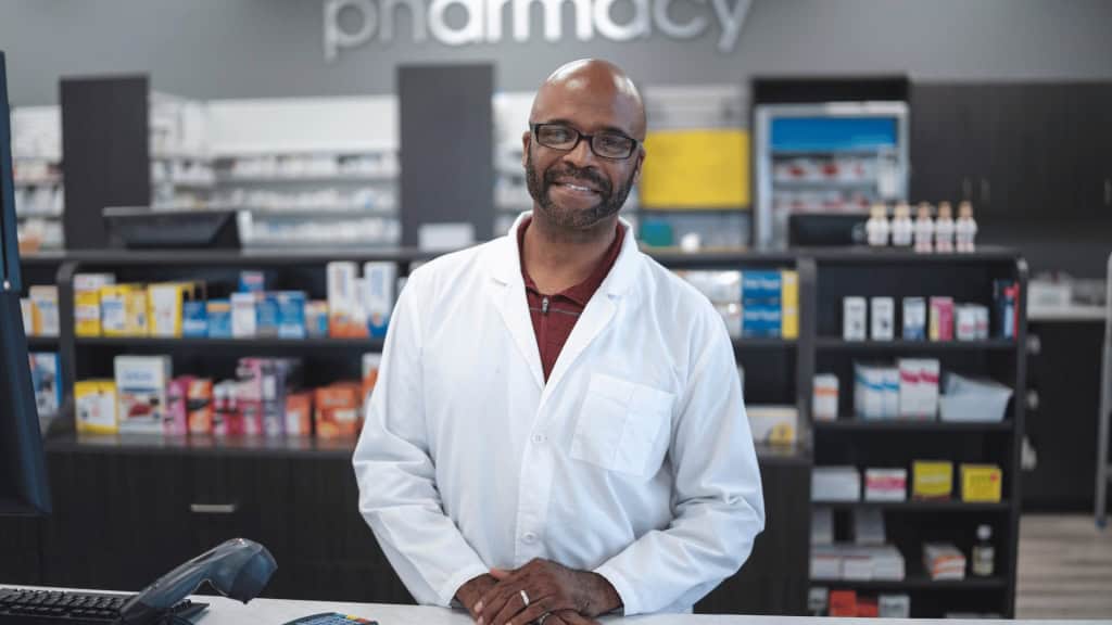 A pharmacist wearing glasses and a white lab coat smiles while standing behind a counter in a pharmacy with shelves of medications in the background.