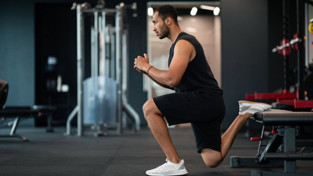 A person is performing a Bulgarian split squat exercise in a gym. Their back foot is elevated on a bench while they lower their front leg into a squat position.
