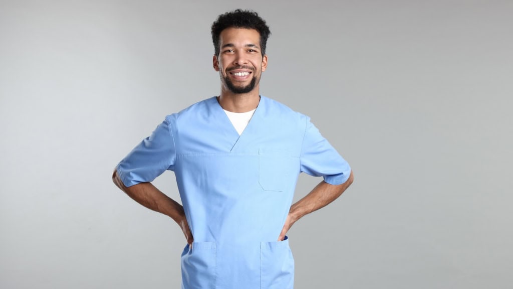 A person wearing a light blue scrub top stands with hands on hips and smiles at the camera against a plain gray background.