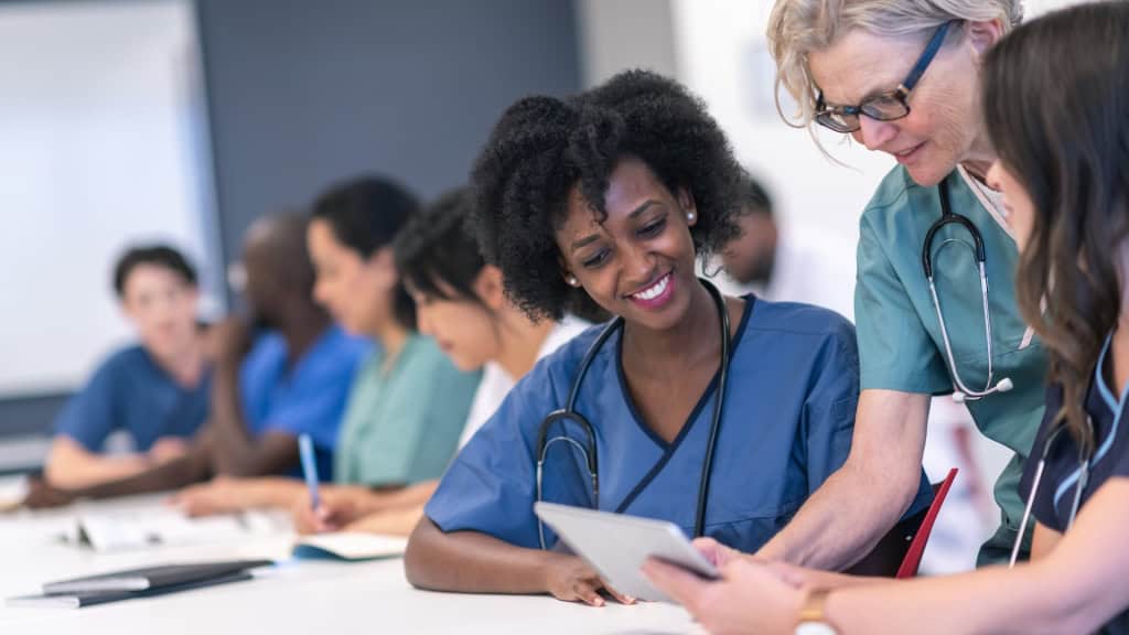 A smiling nursing student in blue scrubs reviews materials with an instructor during a medical class.