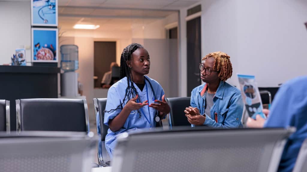 A person in blue scrubs with a stethoscope talks to another person wearing glasses and a denim jacket in a waiting room.