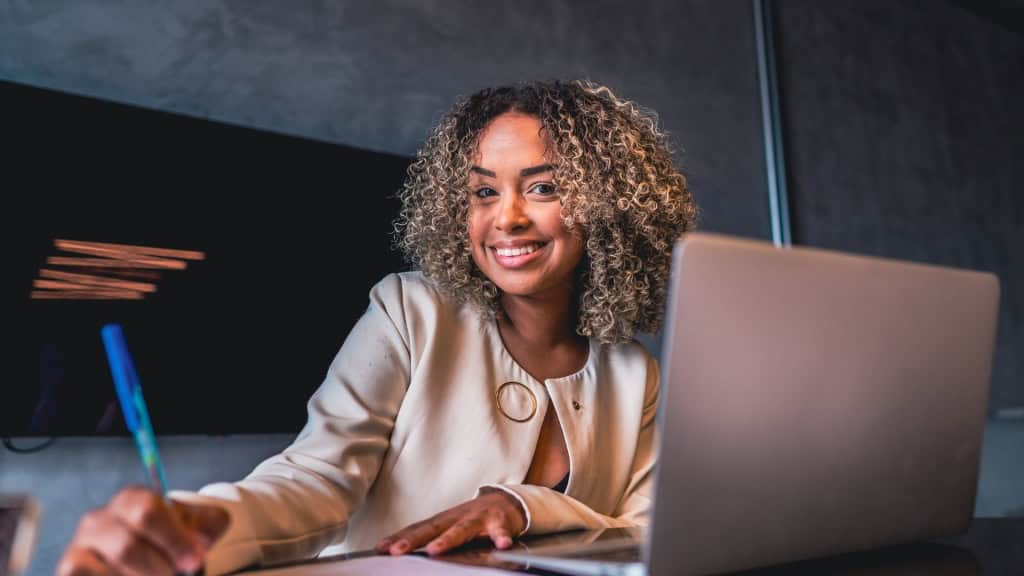 A smiling person with curly hair wearing a light-colored jacket sits at a desk with a laptop and a pen, writing on a document.