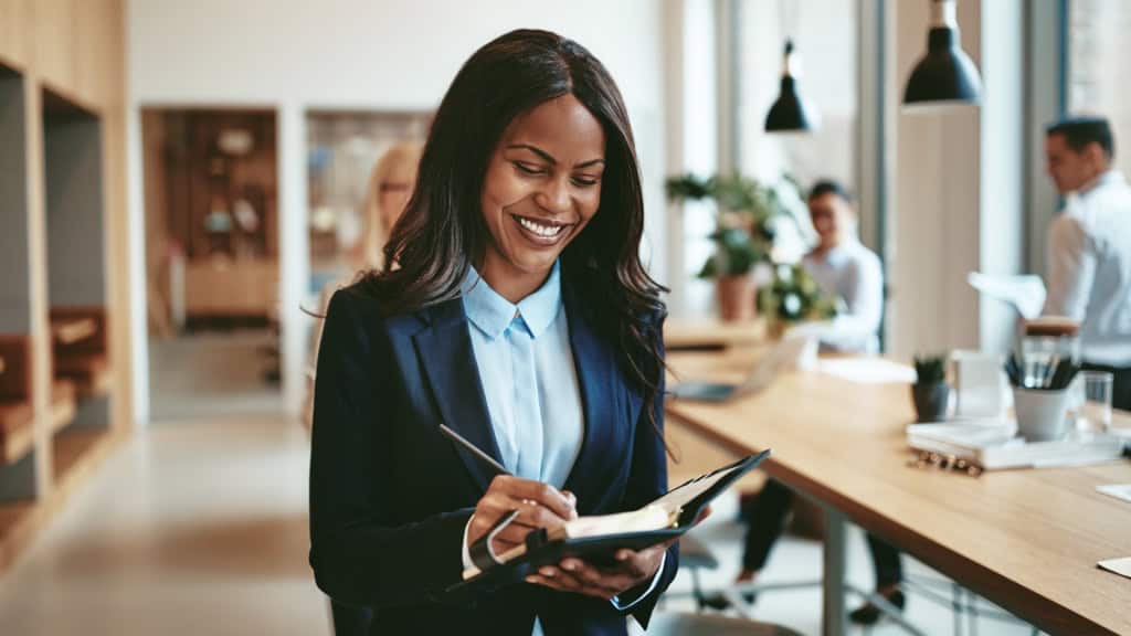 A woman in a suit smiles as she writes in a planner in an office setting with blurred colleagues in the background.