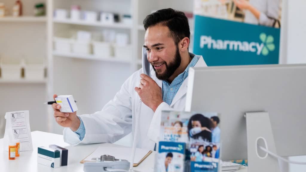 A pharmacist in a white coat holds a medication box while talking on the phone, with a computer and pharmacy shelves in the background.