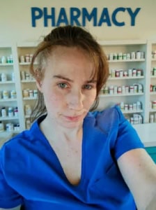 A pharmacist in blue scrubs stands in front of pharmacy shelves stocked with medications.