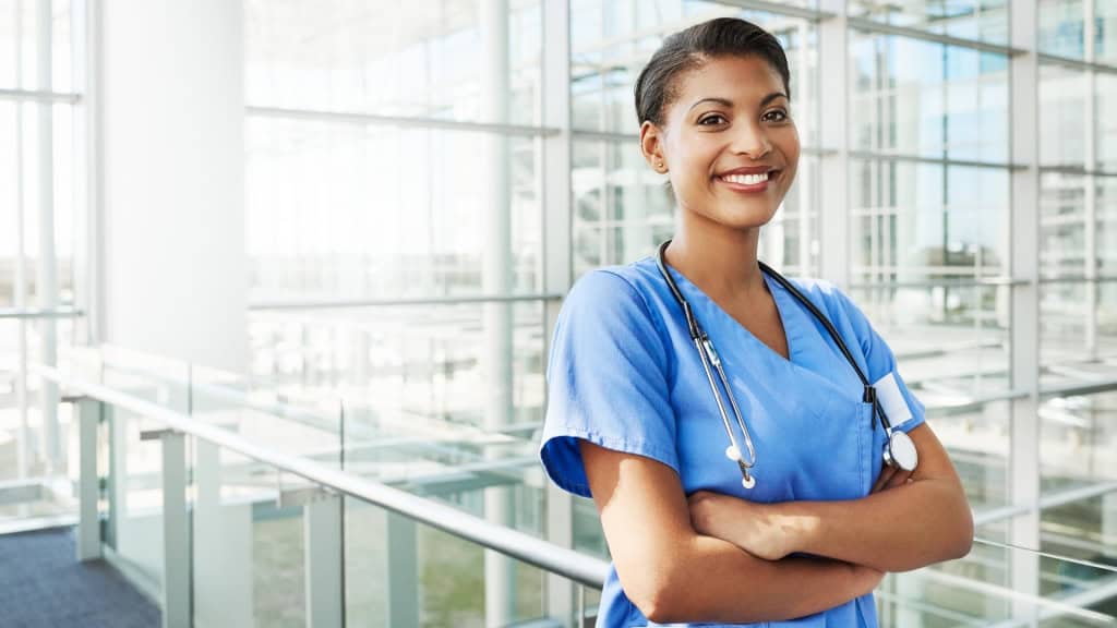 A smiling healthcare professional in blue scrubs with a stethoscope around their neck stands with arms crossed in a modern building with large windows.