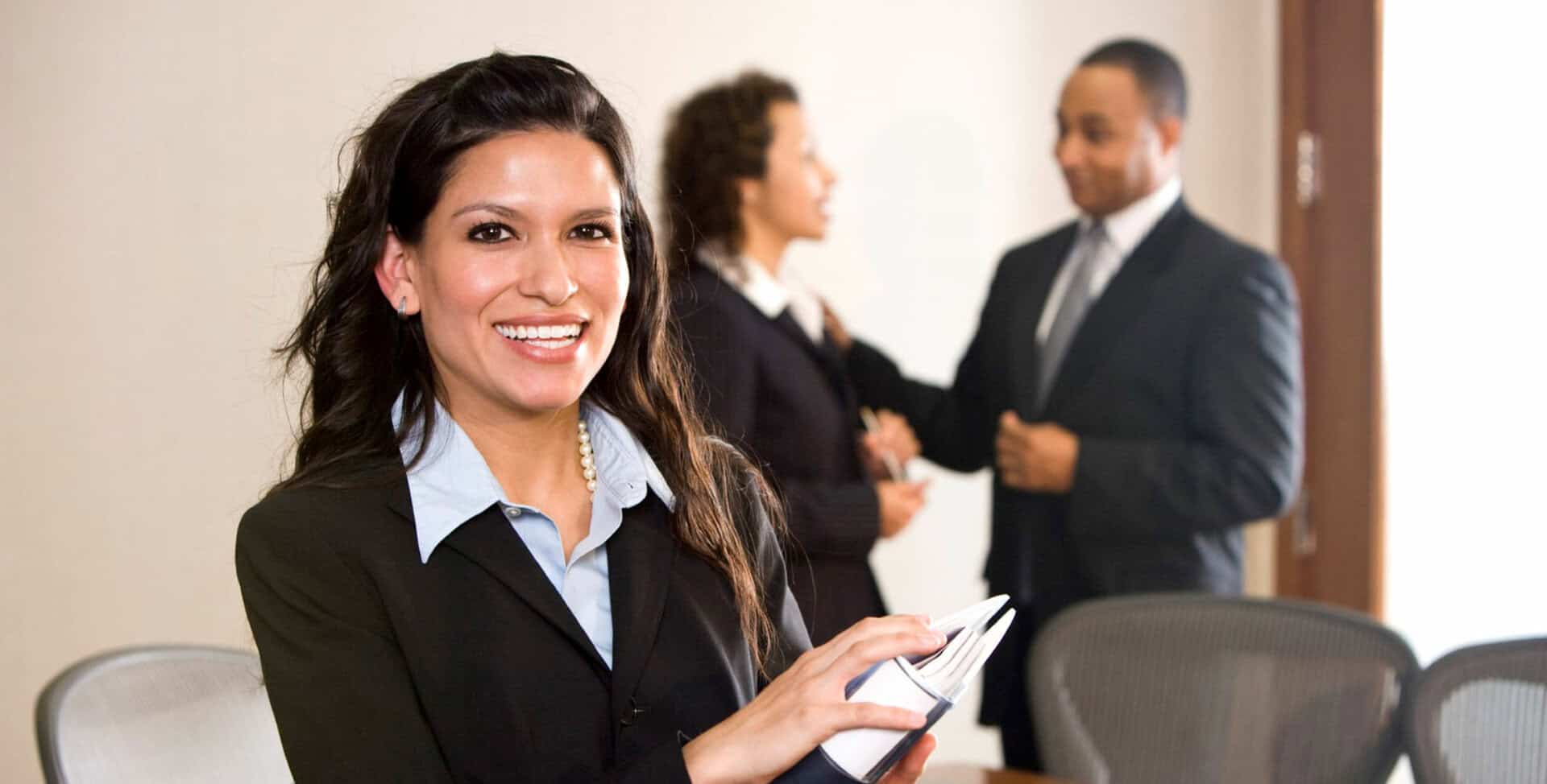 Woman in business suit smiling at camera while holding tablet, with colleagues meeting in background office setting.