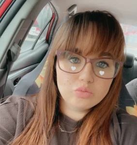 A woman with long reddish-brown hair and glasses taking a selfie while sitting in a car.